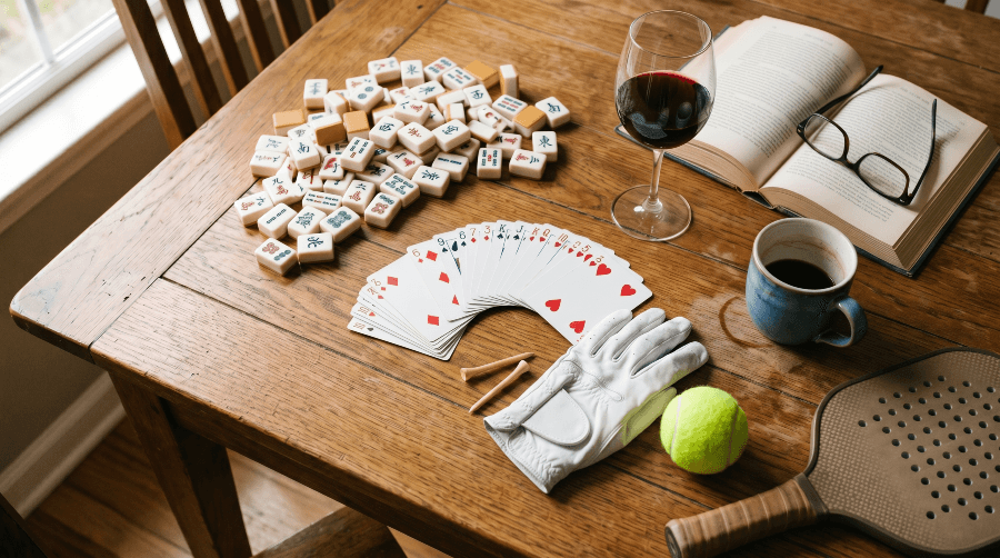A lived-in wooden table casually scattered with the artifacts of one member's club activities: a fanned deck of cards, a loose pile of Mah Jongg tiles, a golf glove and tees, a tennis ball, a glass of wine, and an open book with reading glasses.
