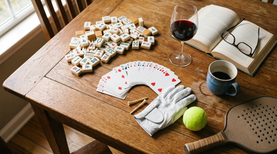 A lived-in wooden table casually scattered with the artifacts of one member's club activities: a fanned deck of cards, a loose pile of Mah Jongg tiles, a golf glove and tees, a tennis ball, a glass of wine, and an open book with reading glasses.